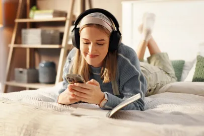 Junge Frau liegt mit Kopfhörern auf dem Bett und nutzt ihr Smartphone